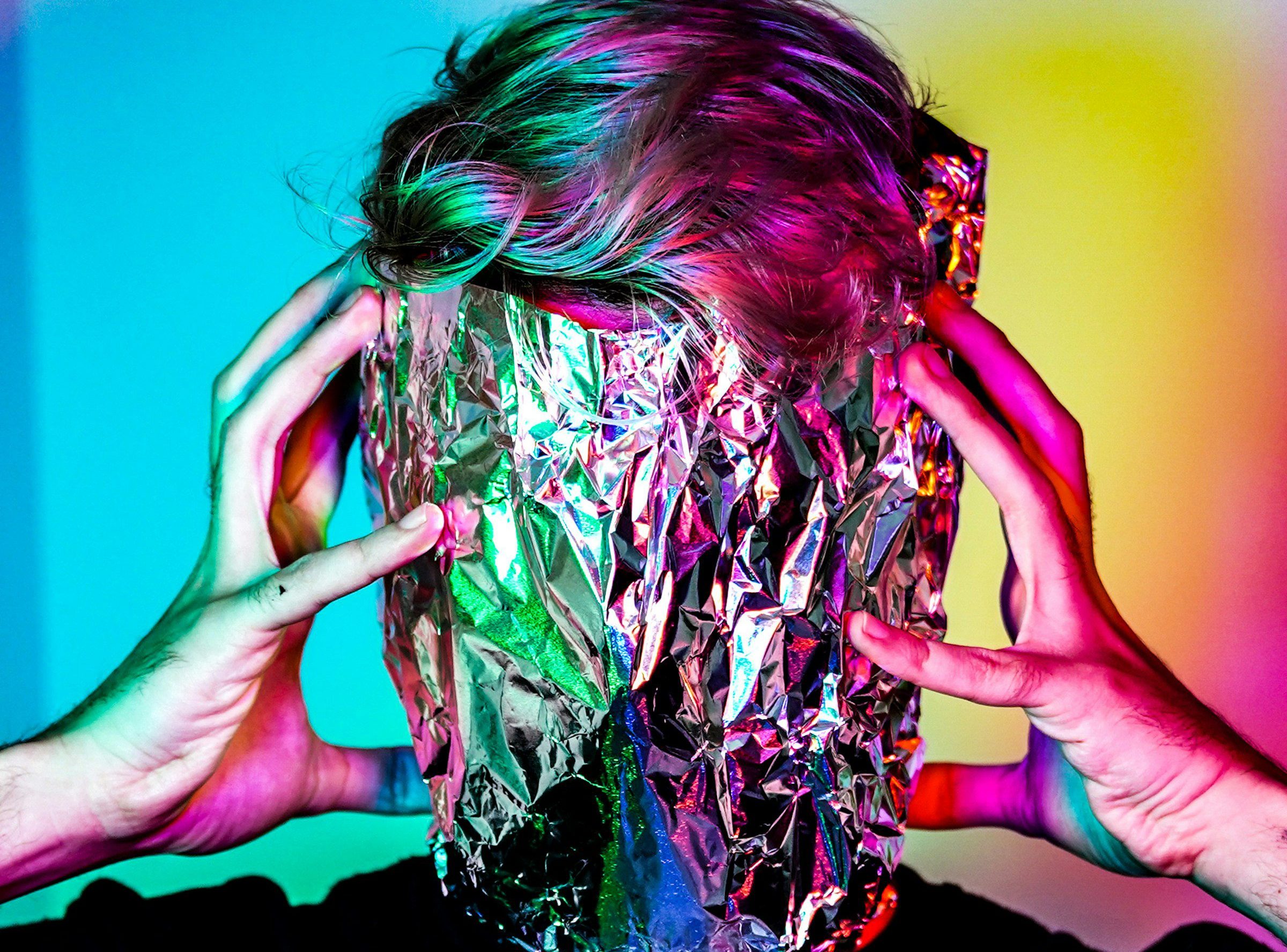 A person with aluminium foil wrapped around their head, surrounded by coloured lights. Image courtesy of Vinicius Amnx Amano (Unsplash)